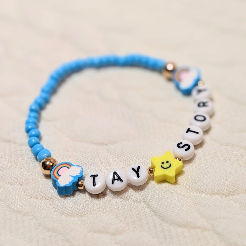 Dainty light blue beads with gold accents rainbows and star in between words that say "Tay Story"