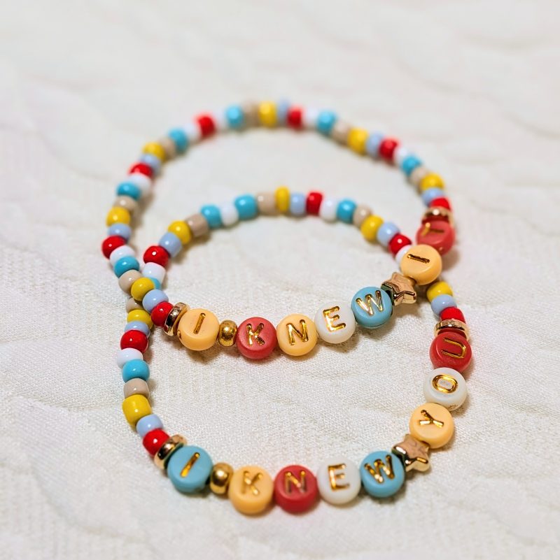 A duo of bracelets. Dainty multicolour beads in tan, red, blue and yellow, with gold accents and letter beads.  One says 