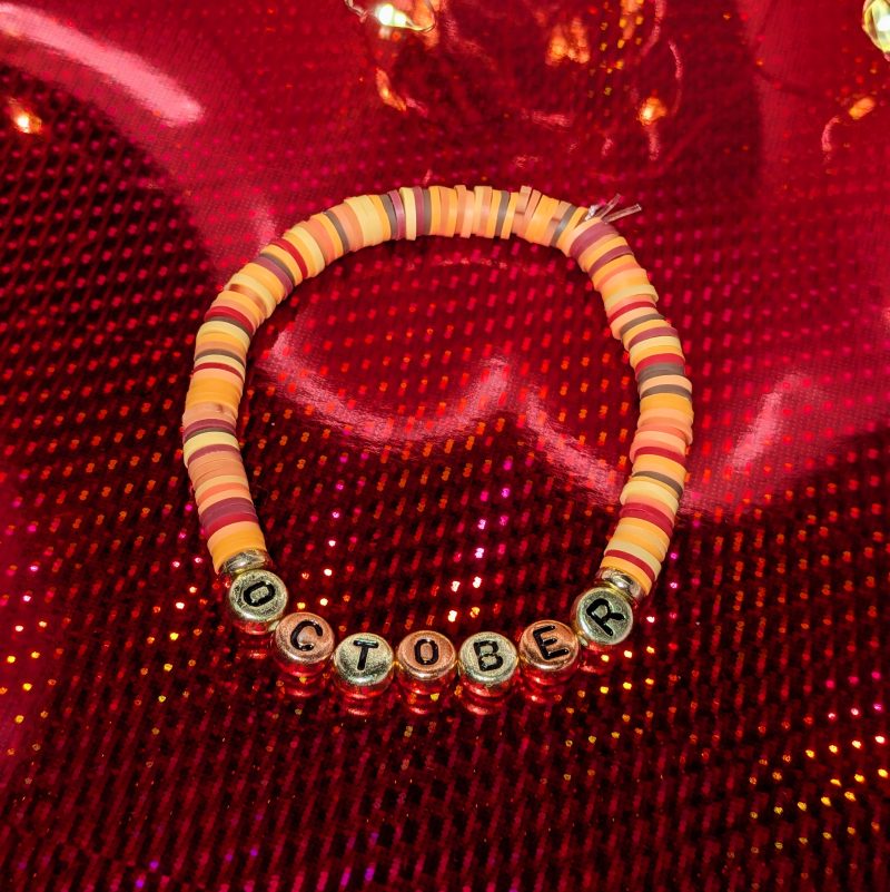 A bracelet that says "October" in gold and bronze letters, with clay beads, and gold spacers — view 2