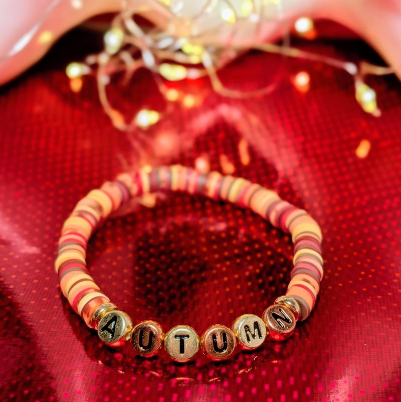 A bracelet that says "Autumn" in gold and bronze letters, with clay beads, and gold spacers