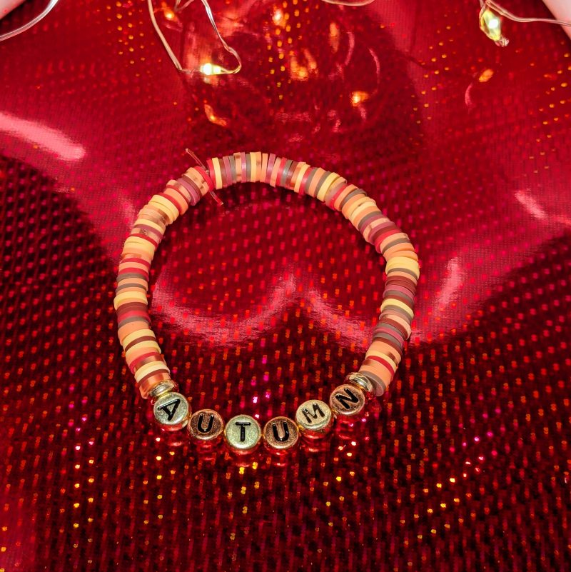 A bracelet that says "Autumn" in gold and bronze letters, with clay beads, and gold spacers — view 2
